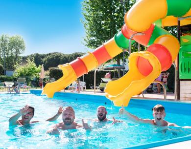 Quattro uomini sorridono in piscina con scivolo colorato.