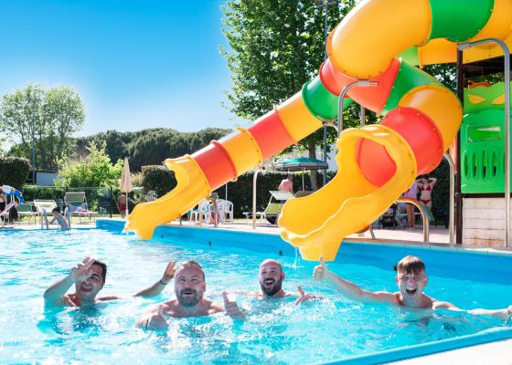 Quattro uomini sorridono in piscina con scivolo colorato.